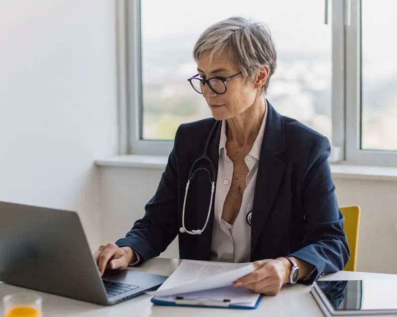 A physician with reading glasses reviewing notes at a laptop by a window